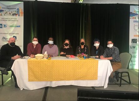 A picture taken of members of Re-Creation Collective during a keynote talk. All of the members are masked and sitting behind a table on a stage. From left to right the members include: William Bridel, Danielle Peers, Janelle Josep, Tricia McGuire-Adams, Cindy Gaudet, Lindsay Eales, Nathan Fawaz.