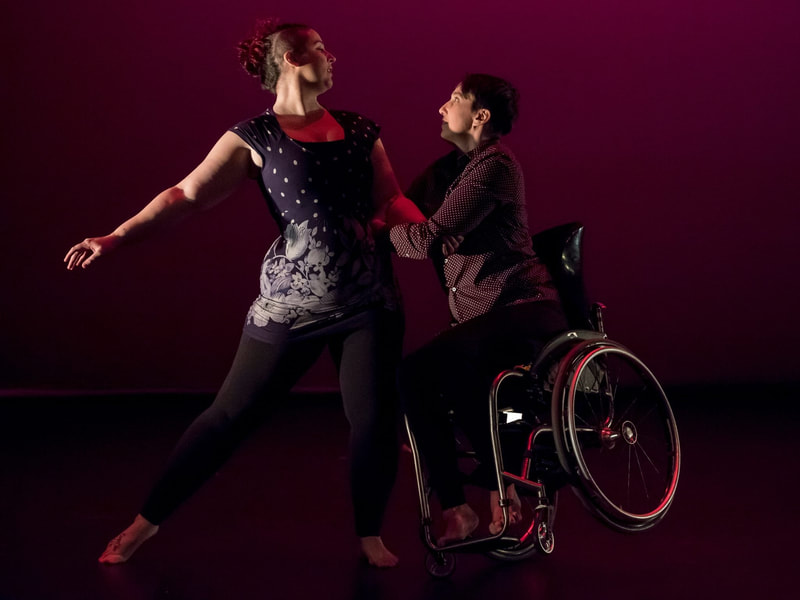 A picture of two people dancing. They are facing each other. One dancer is a chair user, who is balancing on their front wheels while holding the other dancer’s arm. 