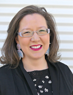 Picture of Tricia. She is smiling in front of a corrugated metal background. She has shoulder length brown hair and glasses. She is wearing beaded earrings, a black and white shirt, and a black blazer.  